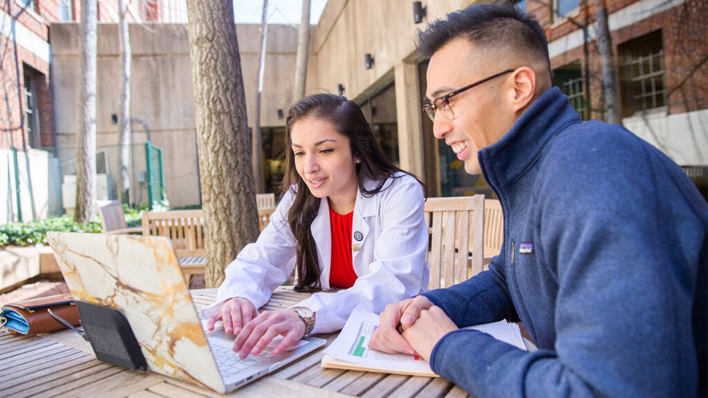 Two medical students work together at a laptop at an outdoor table
